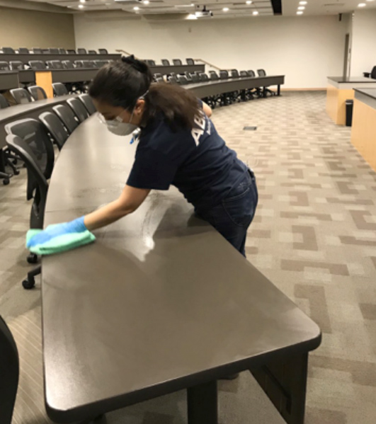 A member of the cleaning crew wears a mask and gloves as she wipes down the surface of a table with sanitizing spray and a towel.