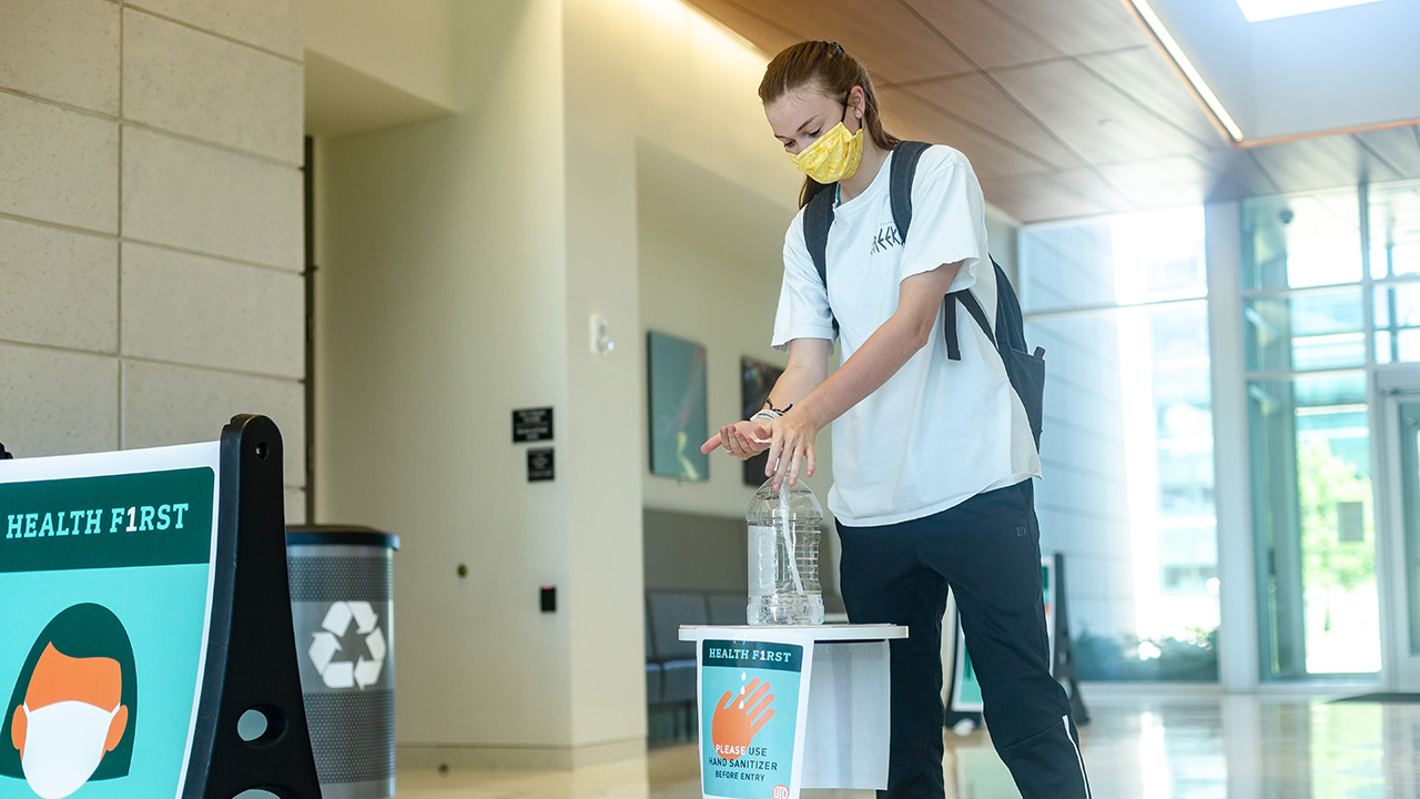 A student uses hand sanitizer at the entrance of a campus building. Signs indicate that people should wear face coverings and use hand sanitizer before proceeding.
