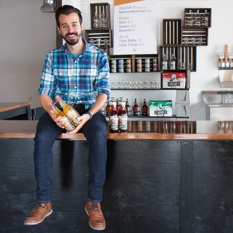 Joel Malone holds bottles of cider as he sits on the bar top at his store.