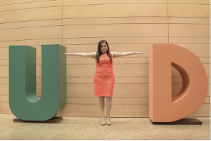 A student holds her arms out to form a T as she stands between a large letter U and D.